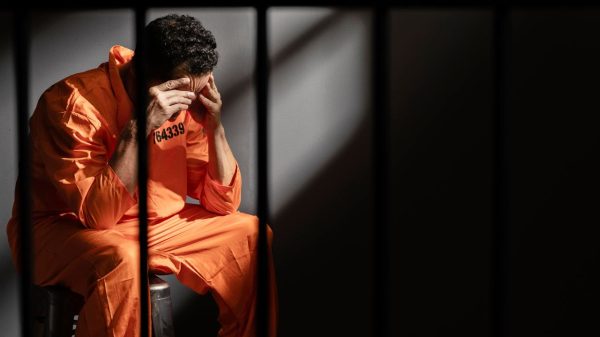 Na imagem um homem preso veste um uniforme laranja de presidiário. Ele expressa sentimento de apreensão pela privação de sua liberdade