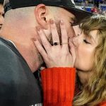 Na imagem há a cantora Taylor Swift e seu namorado o jogador de futebol americano Travis Kelce se beijando em um estádio de futebol. A cantora veste um tricô vermelho e o jogador seu uniforme profissional cinza