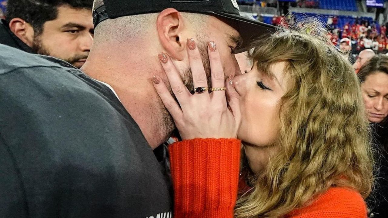 Na imagem há a cantora Taylor Swift e seu namorado o jogador de futebol americano Travis Kelce se beijando em um estádio de futebol. A cantora veste um tricô vermelho e o jogador seu uniforme profissional cinza