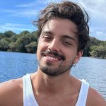 Rodrigo Simas posa sorridente em frente a um lago. Ele veste uma regata branca.