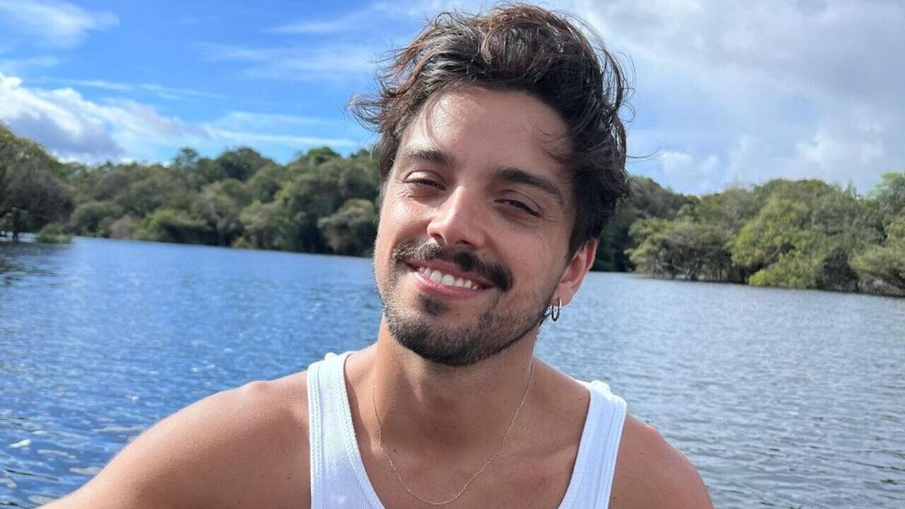 Rodrigo Simas posa sorridente em frente a um lago. Ele veste uma regata branca.