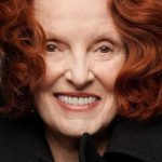 Grace Coddington posa sorridente. Seu cabelo ruivo está escovado e curto. Ela utiliza um batom pêssego com fundo rosado e um casaco preto.