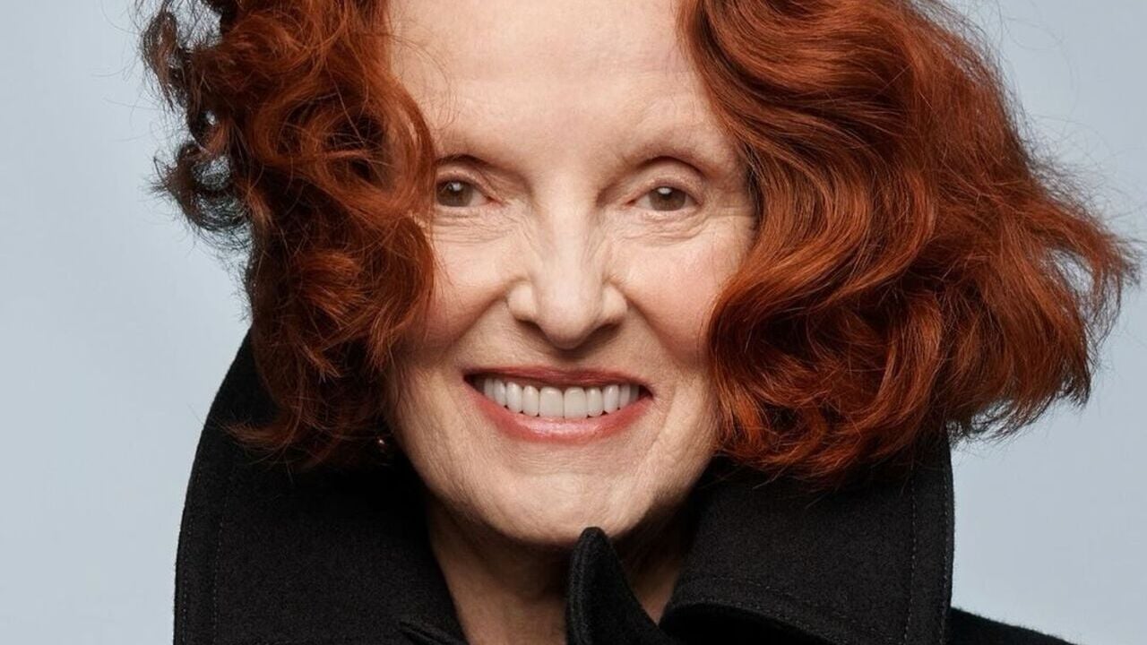 Grace Coddington posa sorridente. Seu cabelo ruivo está escovado e curto. Ela utiliza um batom pêssego com fundo rosado e um casaco preto.