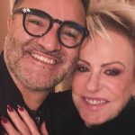 Ana Maria Braga e seu namorado Fabio Arruda posam sorridentes e abraçados. Ambos vestem roupas pretas e Fabio usa um óculos de grau de armação preta.