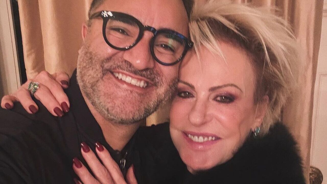 Ana Maria Braga e seu namorado Fabio Arruda posam sorridentes e abraçados. Ambos vestem roupas pretas e Fabio usa um óculos de grau de armação preta.