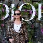 Jennifer Lawrence posa em frente ao letreiro da Dior em desfile num Jardim. Ela veste uma camiseta branca, casaco com estampa de onça e óculos escuros com os cabelos para trás.