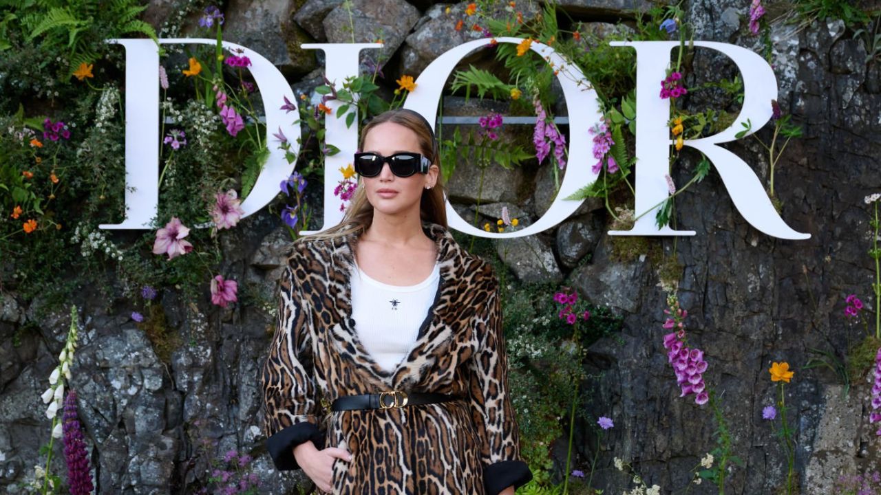 Jennifer Lawrence posa em frente ao letreiro da Dior em desfile num Jardim. Ela veste uma camiseta branca, casaco com estampa de onça e óculos escuros com os cabelos para trás.