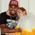 Naldo Benny e Miley Cyrus posam abraçados para foto. Ele veste uma camiseta preta e um boné vermelho. Ela está usando um body amarelo de mangas longas com plumas brancas no ombro.