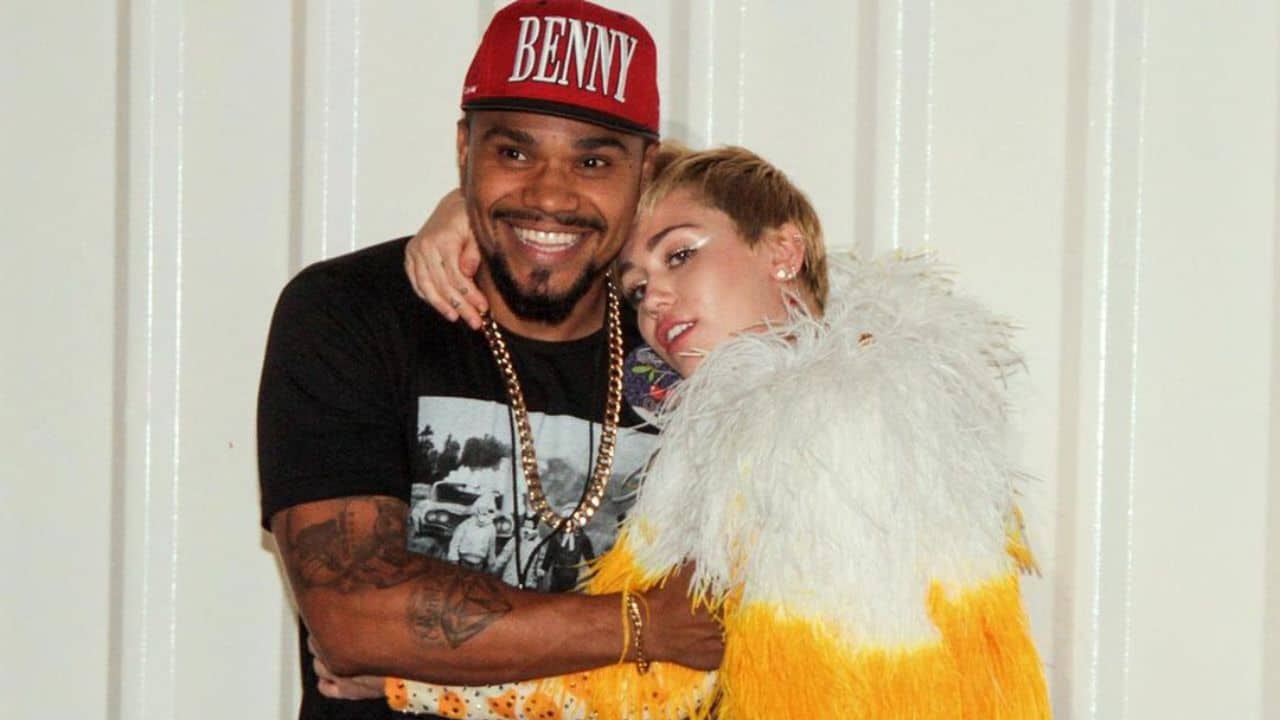 Naldo Benny e Miley Cyrus posam abraçados para foto. Ele veste uma camiseta preta e um boné vermelho. Ela está usando um body amarelo de mangas longas com plumas brancas no ombro.