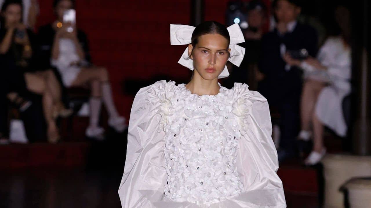 Modelo desfila para Chanel com vestido branco inspirado nos anos 80. A peça tem mangas longas e volumosas, com adornos no torso. O cabelo da modelo está preso para trás e tem como acessório um laço branco.