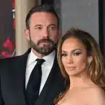 Jennifer Lopez e Ben Affleck durante evento. Ben veste um terno preto e uma camisa branca com gravata preta. Jennifer está com um vestido preto sem mangas.