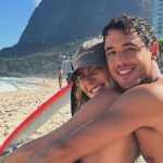 Mariana Goldfarb e Rafael Kemp posam em praia no Rio de Janeiro. Ambos estão usando roupas de banho e sentados sorridentes ao lado de uma prancha de surfe branca. Mariana veste um boné preto.