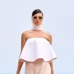 Modelo desfila para Jacquemus. Ela veste um lenço branco que cobre seu cabelo, óculos escuro com armação bege, top geométrico e rodado sem alças com uma saia bege longa e levemente transparente.