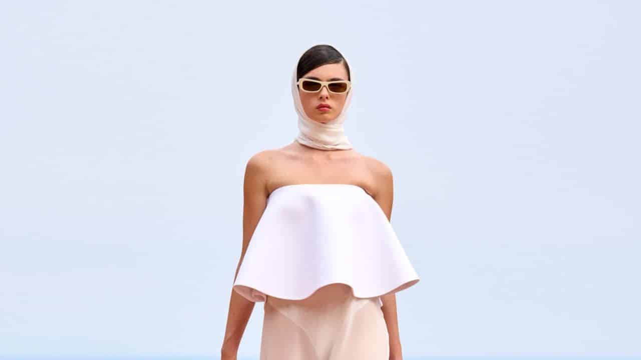 Modelo desfila para Jacquemus. Ela veste um lenço branco que cobre seu cabelo, óculos escuro com armação bege, top geométrico e rodado sem alças com uma saia bege longa e levemente transparente.