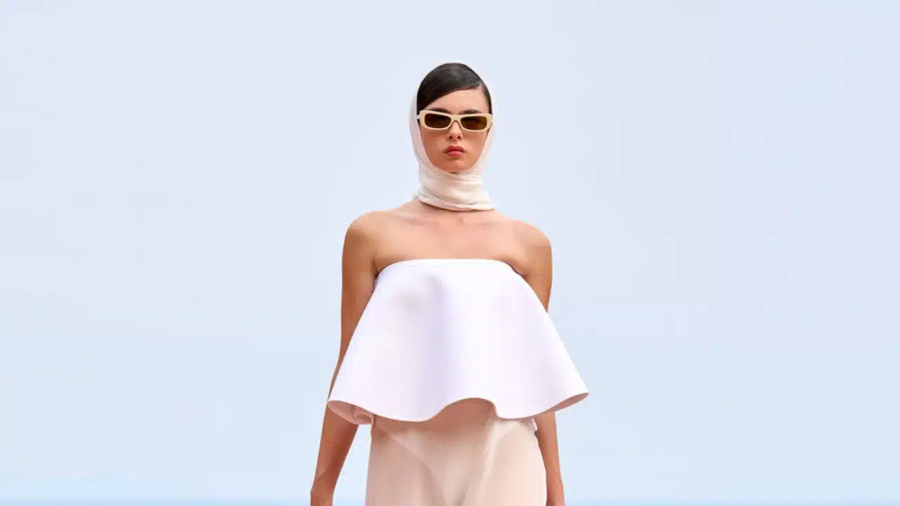 Modelo desfila para Jacquemus. Ela veste um lenço branco que cobre seu cabelo, óculos escuro com armação bege, top geométrico e rodado sem alças com uma saia bege longa e levemente transparente.