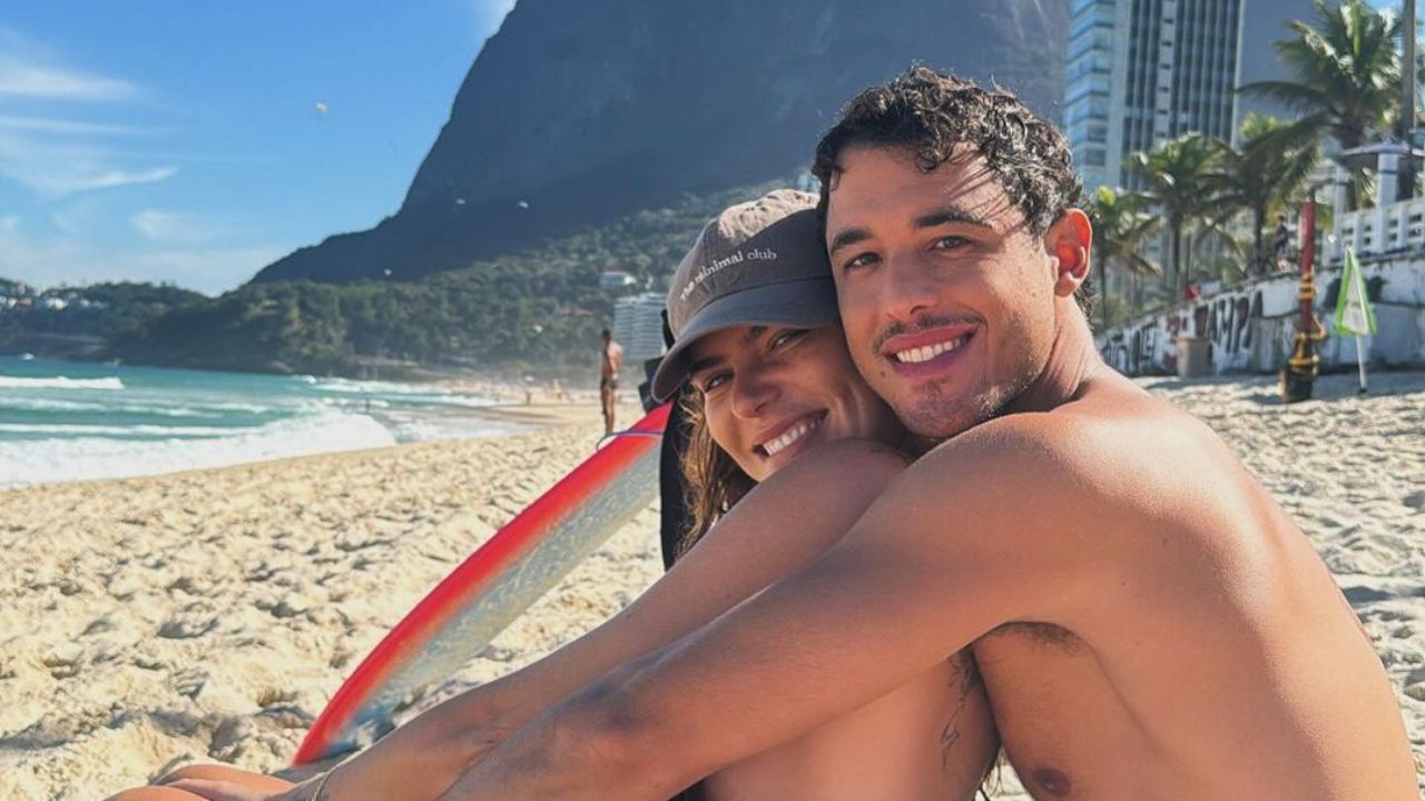 Mariana Goldfarb e Rafael Kemp posam em praia no Rio de Janeiro. Ambos estão usando roupas de banho e sentados sorridentes ao lado de uma prancha de surfe branca. Mariana veste um boné preto.