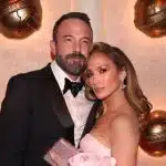 Ben Affleck e Jennifer Lopez posam em frente a parede iluminada com globos de ouro. Ele veste um terno preto e ela um vestido rosa sem alças.