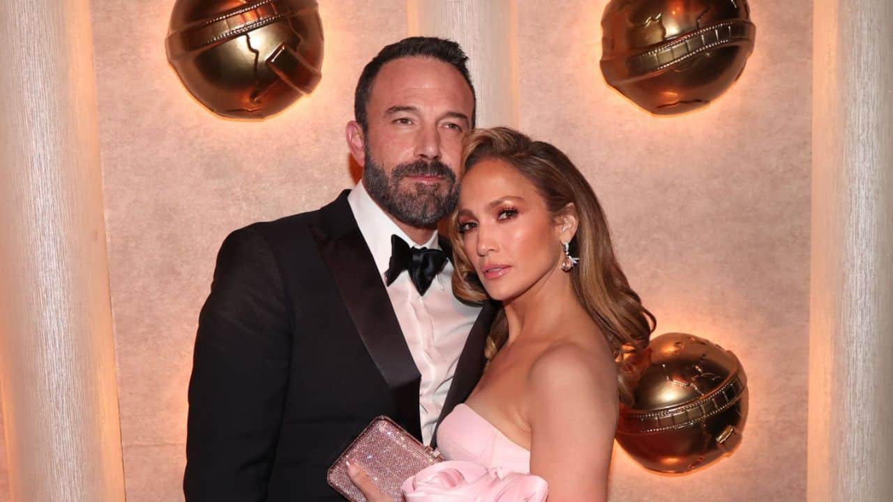 Ben Affleck e Jennifer Lopez posam em frente a parede iluminada com globos de ouro. Ele veste um terno preto e ela um vestido rosa sem alças.