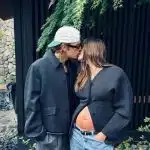 Justin e Hailey Bieber posam se beijando em frente a jardim. Ele veste um boné branco e um casaco preto. A modelo está com um blazer preto aberto mostrando a barriga de grávida e calça jeans.