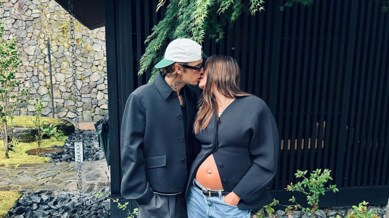 Justin e Hailey Bieber posam se beijando em frente a jardim. Ele veste um boné branco e um casaco preto. A modelo está com um blazer preto aberto mostrando a barriga de grávida e calça jeans.