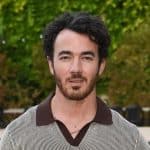 Cantor Kevin Jonas posa em frente a jardim. Ele veste uma camisa listada branca e marrom