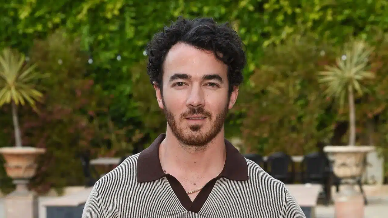 Cantor Kevin Jonas posa em frente a jardim. Ele veste uma camisa listada branca e marrom