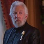 Donald Sutherland como presidente Snow, veste roupas pretas e tem barba e cabelos brancos. Seu rosto e metade do peitoral estão perto da câmera, e o cenário é uma sala com janela e cortina.