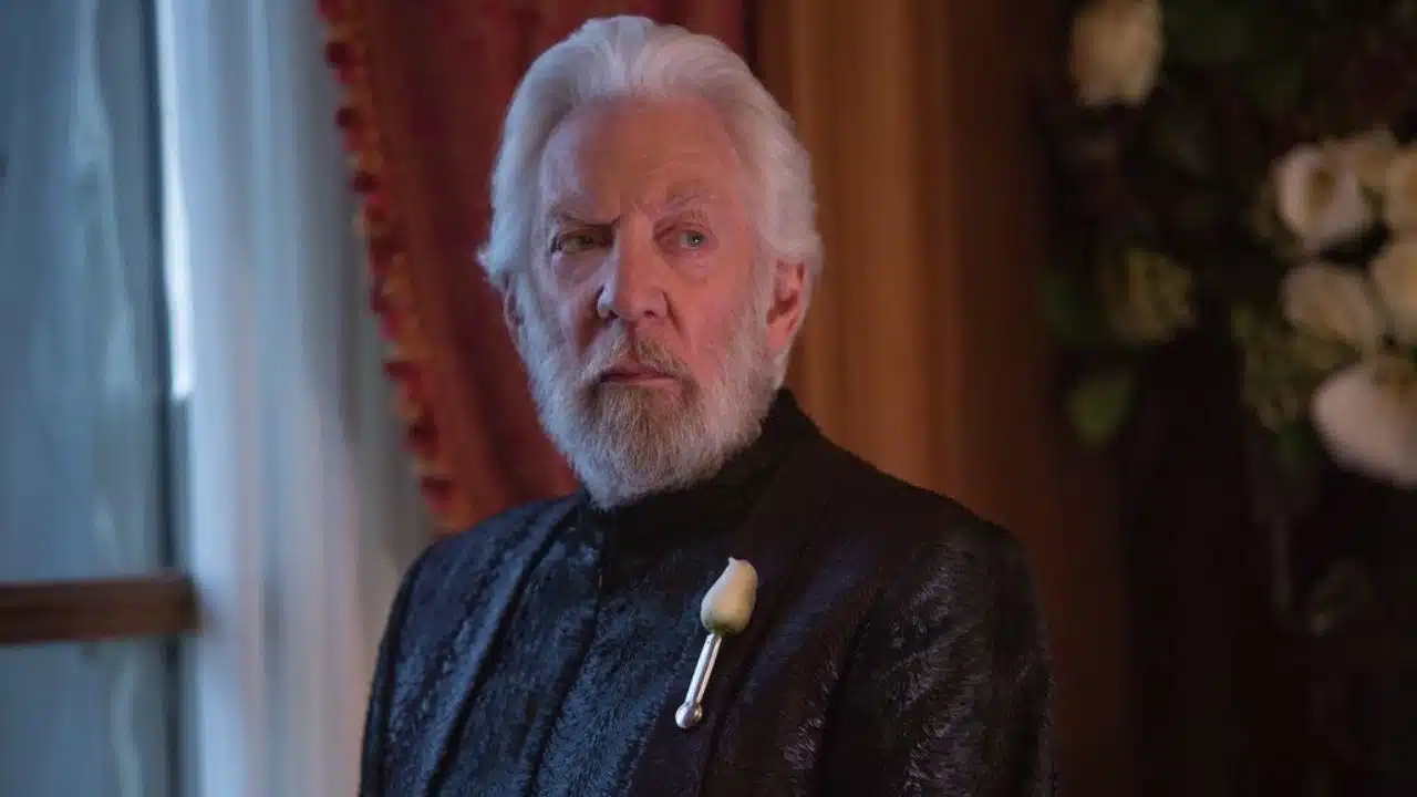 Donald Sutherland como presidente Snow, veste roupas pretas e tem barba e cabelos brancos. Seu rosto e metade do peitoral estão perto da câmera, e o cenário é uma sala com janela e cortina.