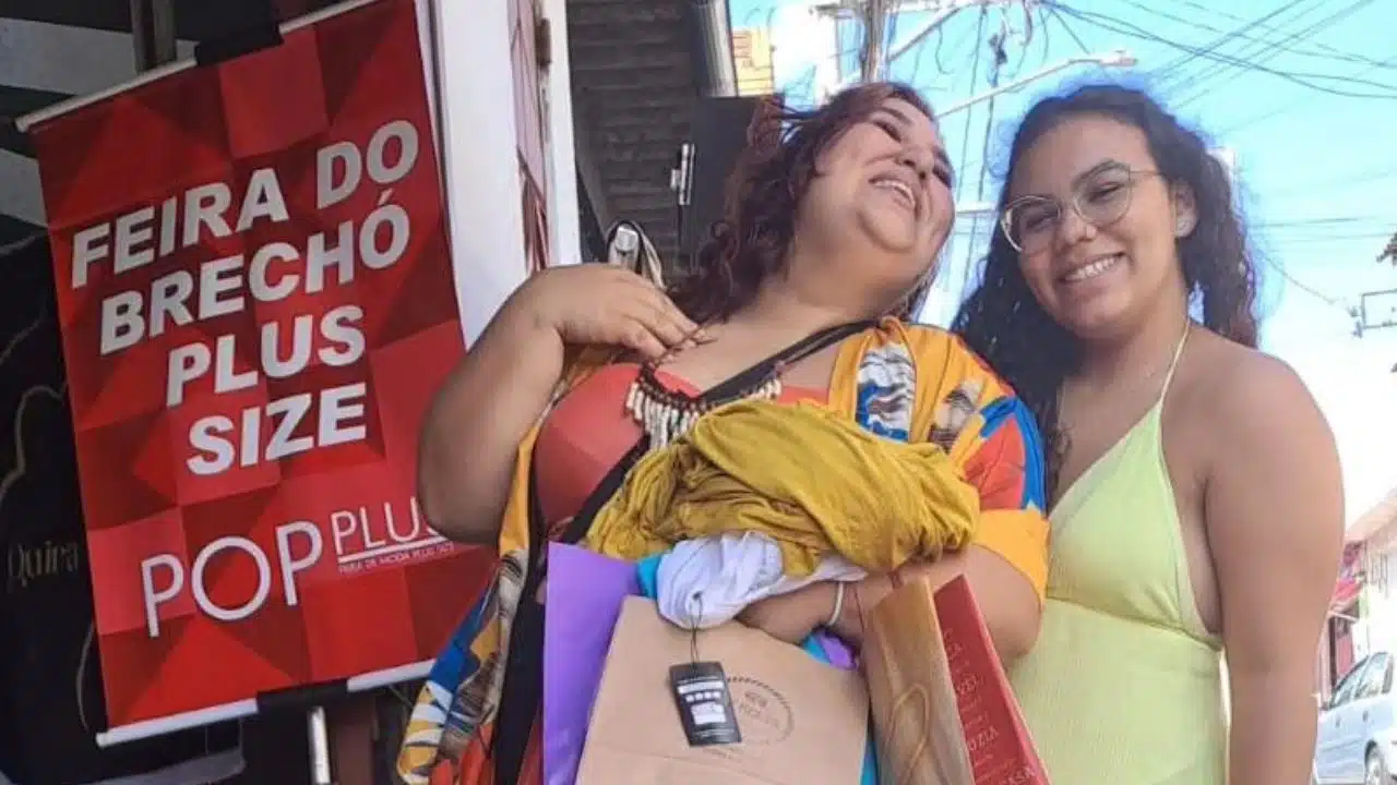 Duas mulheres posam para uma placa vermelha escrita "Feira do brechó plus size POPlus"