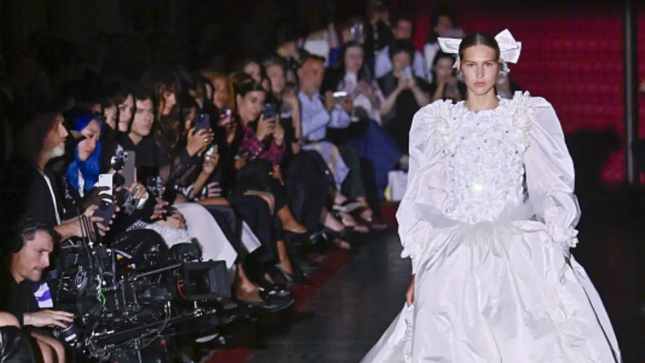 Angelina Kendall nas passarelas vestida de noiva, ela está do lado direito, e no lado esquerdo estão pessoas assistindo ao desfile e pessoas tirando fotos.