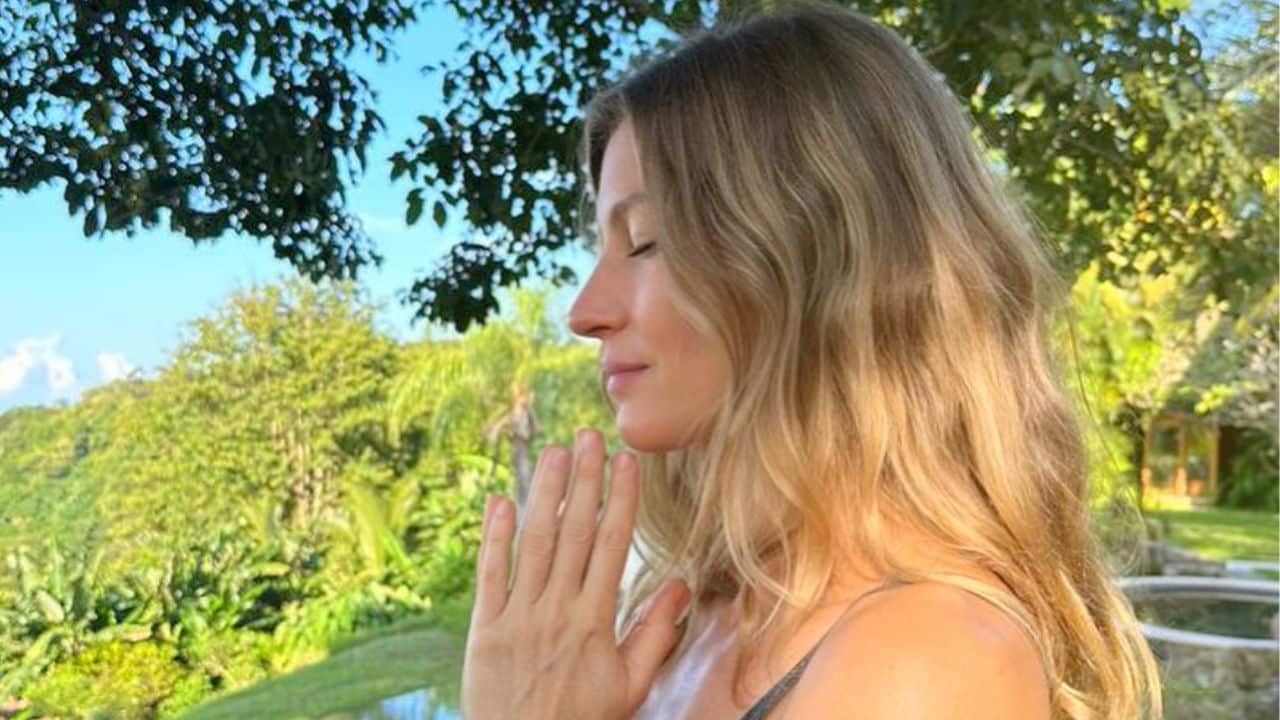 Gisele Bündchen posa com duas mãos em gesto de oração. De fundo, a paisagem é de mato.