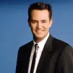 Na imagem há o ator Matthew Perry, ele veste terno risca de giz preto, camisa branca e gravada marrom. Sorri, olhando diretamente para a câmera.