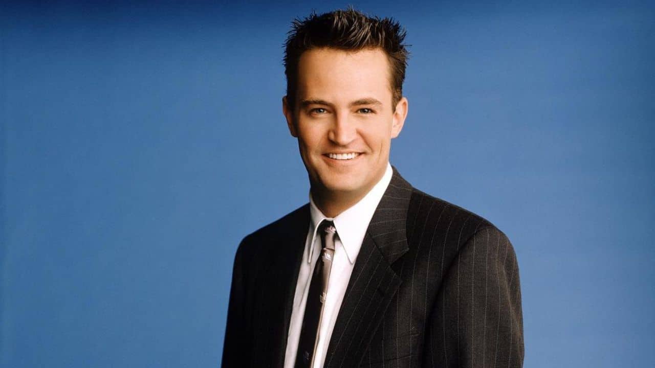 Na imagem há o ator Matthew Perry, ele veste terno risca de giz preto, camisa branca e gravada marrom. Sorri, olhando diretamente para a câmera.