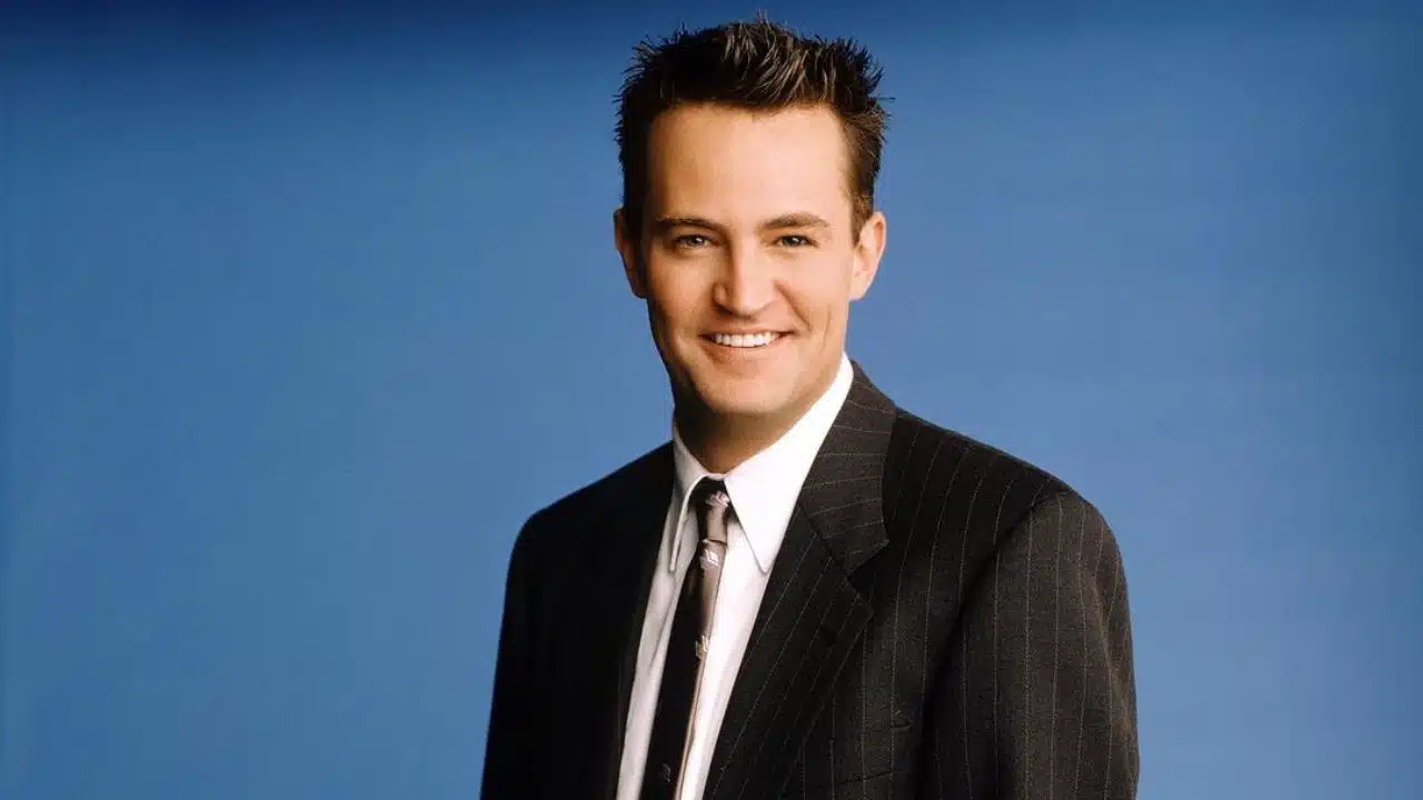 Na imagem há o ator Matthew Perry, ele veste terno risca de giz preto, camisa branca e gravada marrom. Sorri, olhando diretamente para a câmera.