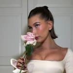 Hailey Bieber com vestido branco, cabelo amarrado para trás e uma flor rosa em suas mãos.