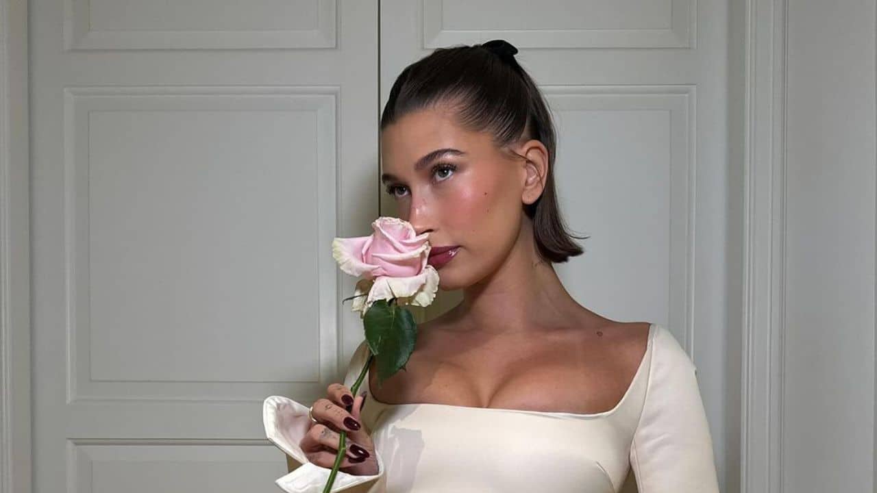 Hailey Bieber com vestido branco, cabelo amarrado para trás e uma flor rosa em suas mãos.