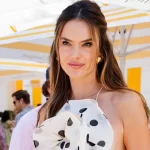 Alessandra Ambrósio posando em um evento ao ar livre, usando uma blusa branca com estampas de bolinha pretas e sorrindo para câmera