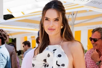 Alessandra Ambrósio posando em um evento ao ar livre, usando uma blusa branca com estampas de bolinha pretas e sorrindo para câmera