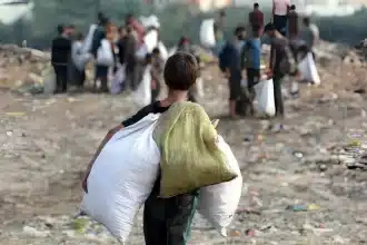 Uma criança de costas carregando dois sacos grandes enquanto caminha por uma área cheia de entulho