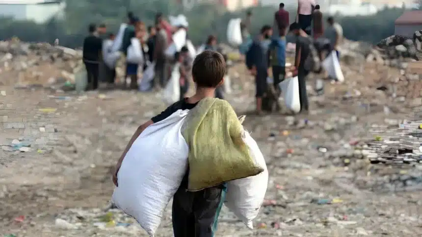 Uma criança de costas carregando dois sacos grandes enquanto caminha por uma área cheia de entulho