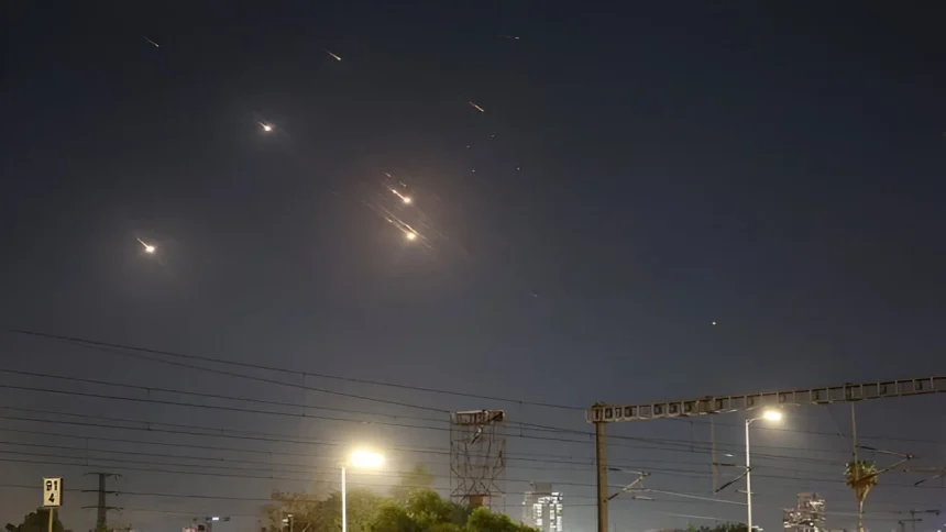 Mísseis iluminam o céu noturno sobre Tel Aviv, com várias explosões visíveis, enquanto o sistema de defesa antimísseis tenta interceptá-los