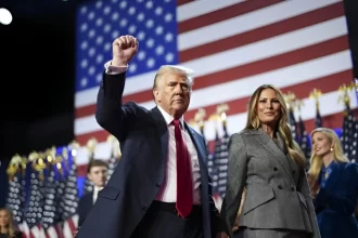 Donald Trump, ao lado de sua esposa Melania Trump, ergue o punho em sinal de vitória, com a bandeira dos Estados Unidos ao fundo, durante o discurso de celebração por sua reeleição à presidência