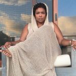 Lizzo sentada, vestindo um vestido longo de crochê com capuz da marca brasileira Ateliê Mão de Mãe.