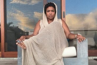 Lizzo sentada, vestindo um vestido longo de crochê com capuz da marca brasileira Ateliê Mão de Mãe.