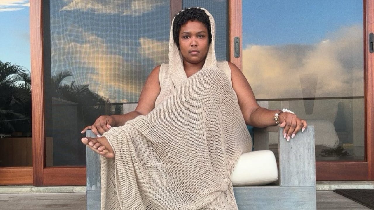 Lizzo sentada, vestindo um vestido longo de crochê com capuz da marca brasileira Ateliê Mão de Mãe.