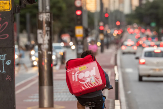 Entregador de aplicativo pedalando em ciclovia movimentada em São Paulo, com mochila térmica do iFood