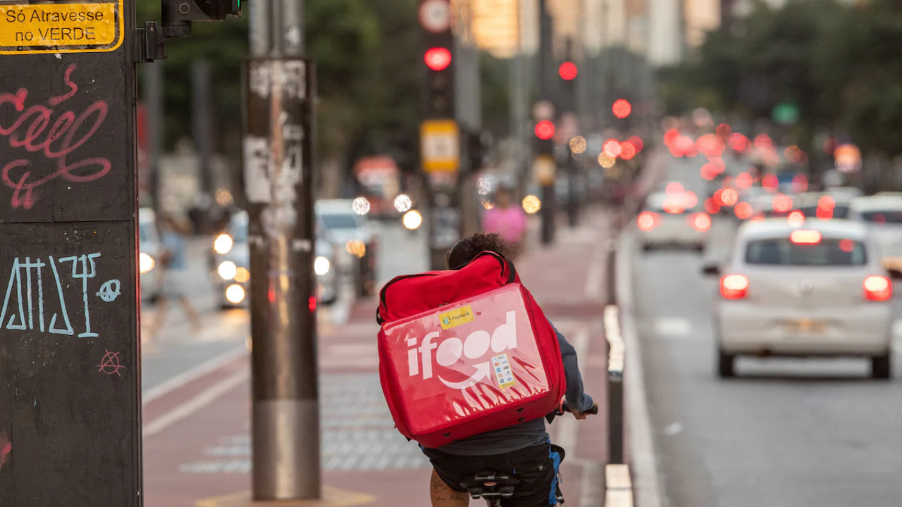 Entregador de aplicativo pedalando em ciclovia movimentada em São Paulo, com mochila térmica do iFood