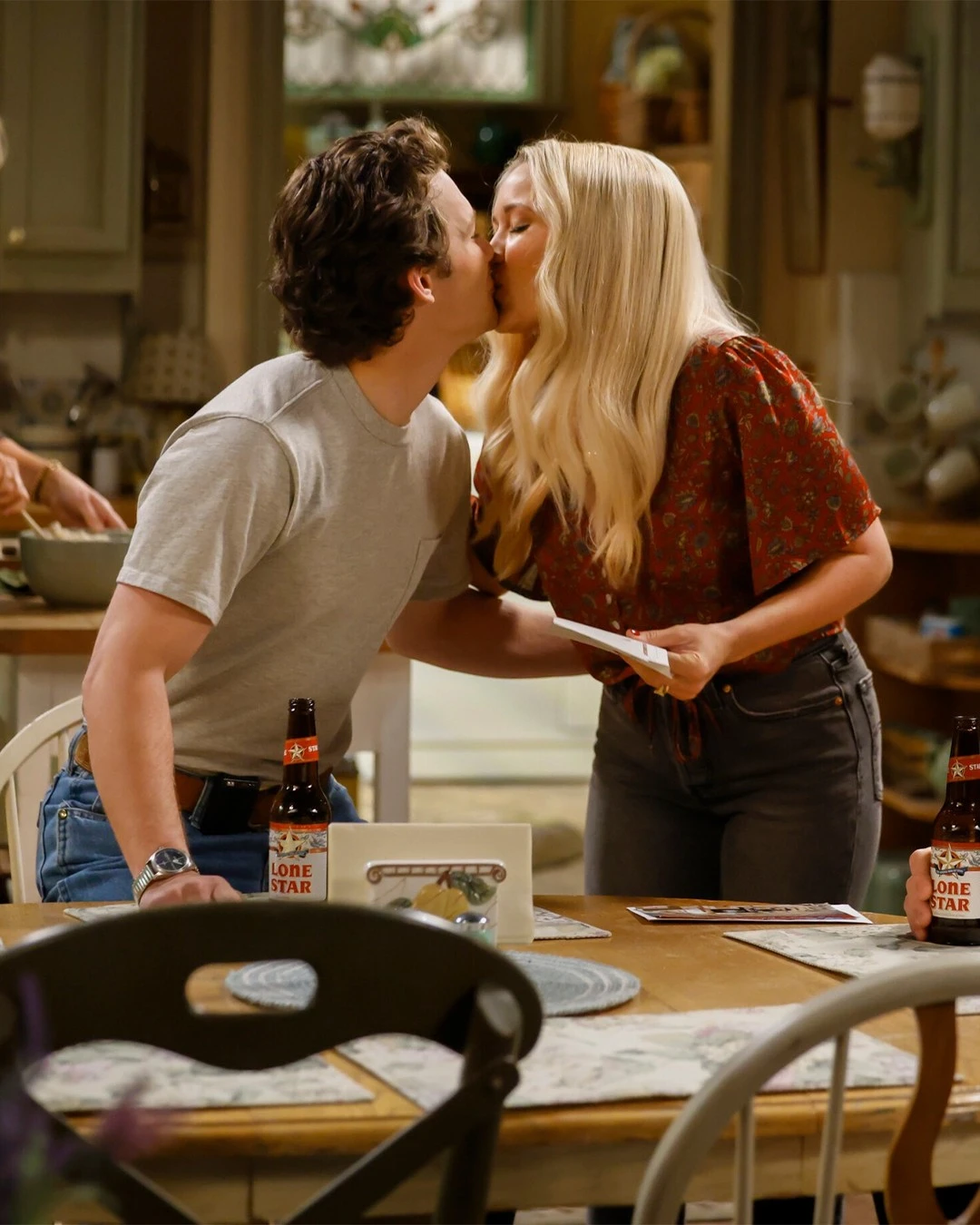 
Georgie e Mandy  se beijam na cozinha enquanto estão ao redor de uma mesa com garrafas de cerveja Lone Star, em um ambiente doméstico e acolhedor.