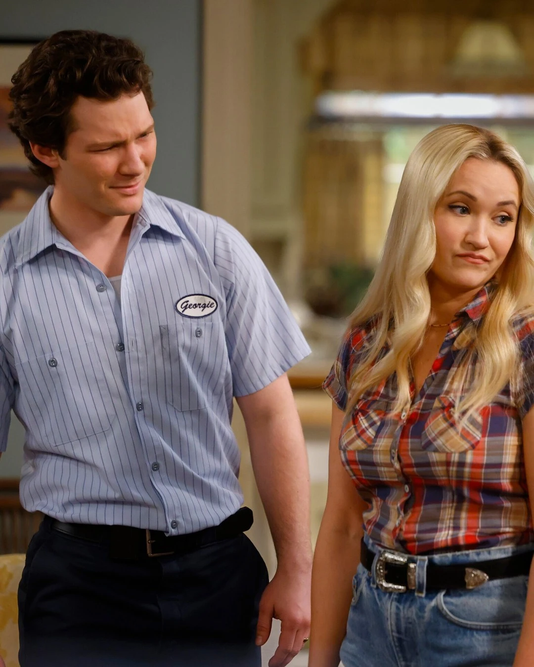 Georgie e Mandy em uma cena doméstica. Georgie veste uniforme de trabalho e olha confuso para Mandy, que responde com uma expressão cética, usando camisa xadrez e shorts jeans.