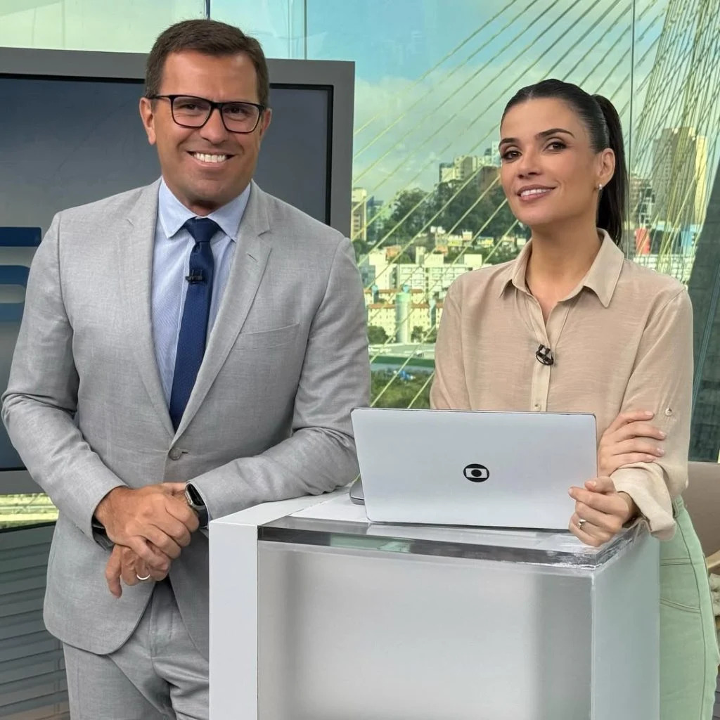 Fundo telejornal os apresentadores olhando para frente em cima da mesa um computaor Simone segurando